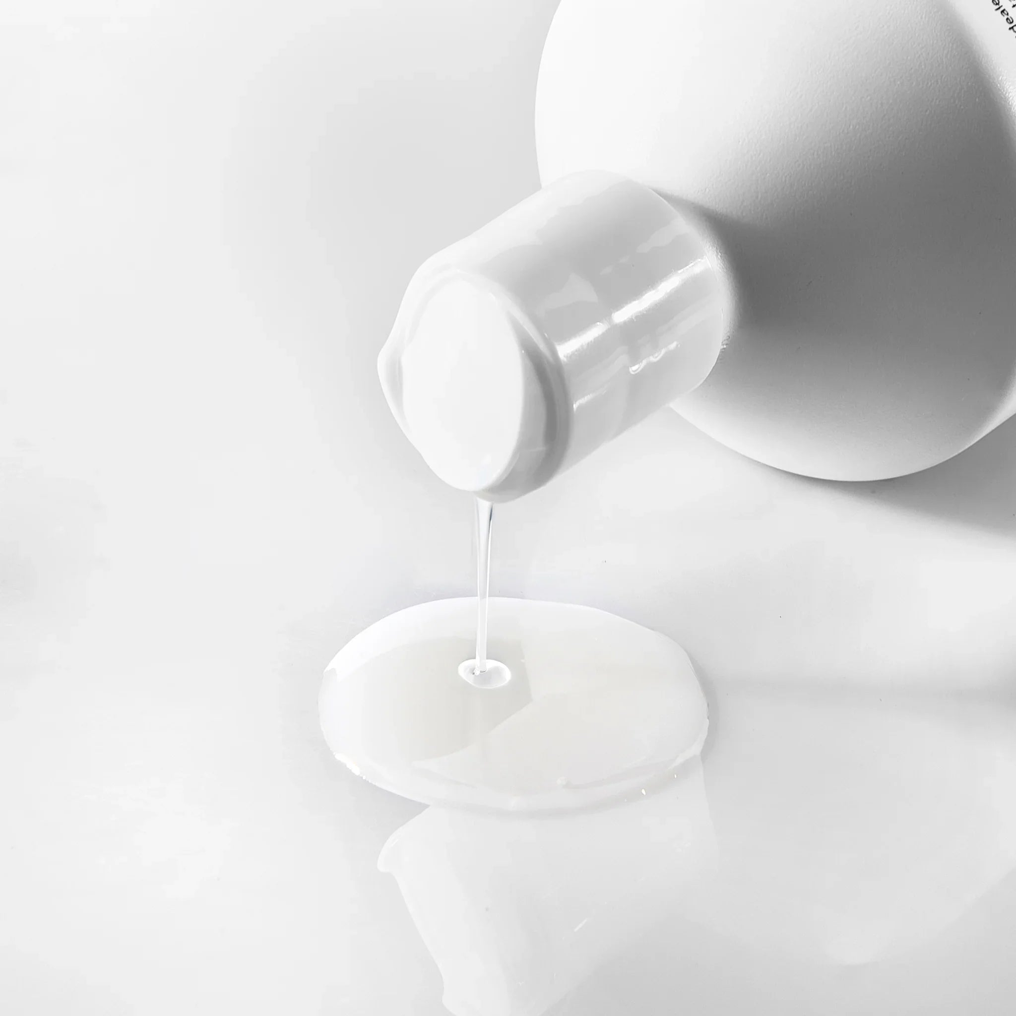 Flacone bianco che versa liquido trasparente su superficie bianca, bagnodoccia delicato